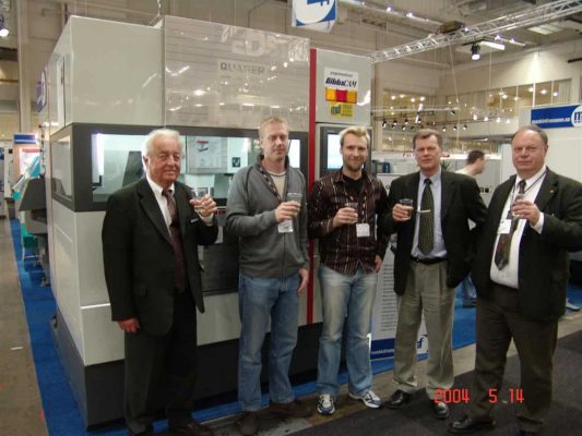 Fem män som står framför en tillverkningsmaskin på en mässa och håller i drinkar, daterad den 14 maj 2004.
