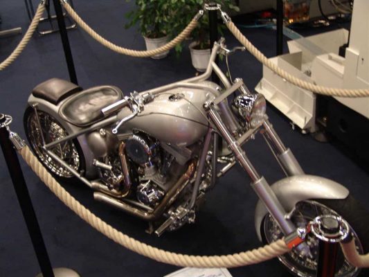 En silver anpassad chopper motorcykel visas på ett heltäckningsmatta golv, omgiven av rep barriärer. Det finns växter i bakgrunden.