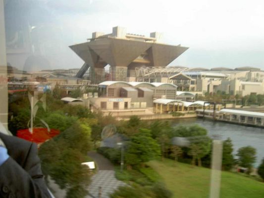 Avlägsen vy av en stor, futuristisk byggnad med en distinkt omvänd pyramidform, omgiven av grönska och mindre strukturer, belägen intill en vattenmassa.