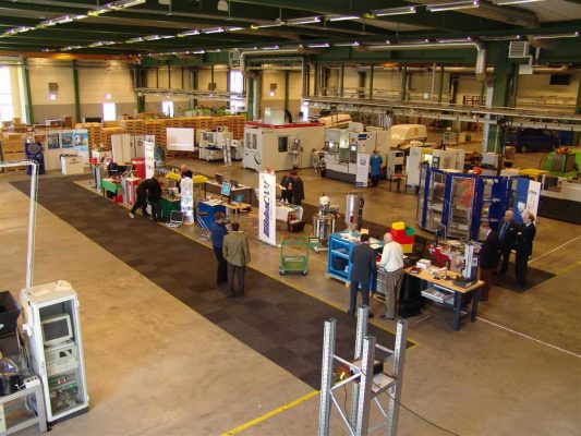 En rymlig industriverkstad med olika maskiner och arbetsstationer. Flera personer är engagerade i olika aktiviteter, undersöker utrustning och samtalar. Området är rent och välorganiserat.