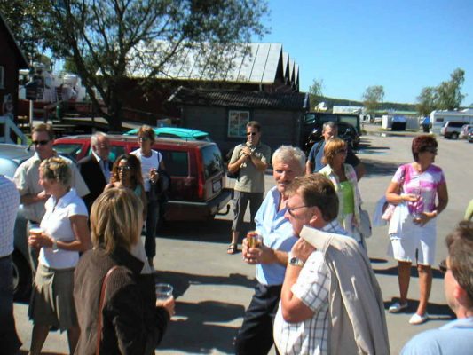 En grupp människor står och samtalar på en parkeringsplats en solig dag, med bilar och träd synliga i bakgrunden. Flera personer håller drinkar.