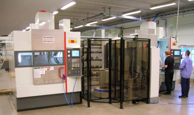 En stor CNC-maskin i en väl upplyst industriverkstad, med två personer stående bredvid, engagerade sig i diskussion.