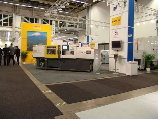 En gul FANUC-utställningsmonter som visar industrimaskiner på en mässa, med ytterligare utrustning och besökare synliga i bakgrunden.