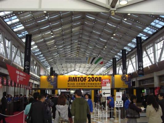 Fullsatt utställningshall med människor som går och en stor banderoll med texten "JIMTOF 2008" hängande under ett trekantigt glastak.