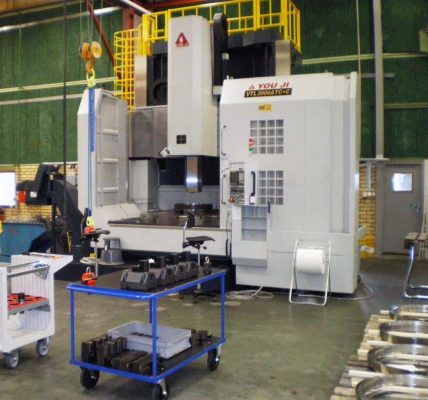 En stor industriell CNC-maskin med kontrollpaneler och en spindel står i fabriksmiljö, omgiven av olika metalldelar, vagnar och verktyg på golvet.