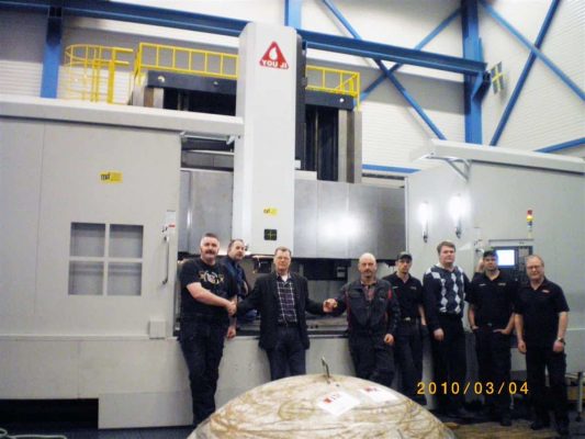 En grupp på åtta män står framför en stor industrimaskin i en fabriksmiljö. Maskinen har en vit och gul struktur. Datumet på bilden är 2010/03/04.