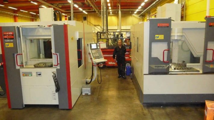 En person står mellan två stora industriella CNC-maskiner i en fabriksmiljö. Maskinerna har kontrollpaneler med olika knappar och displayer.
