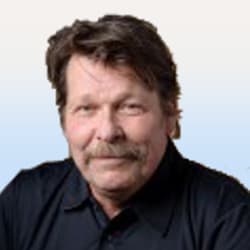 En man med brunt hår, mustasch och ett litet leende, klädd i en svart skjorta, mot en ljus gradientbakgrund.