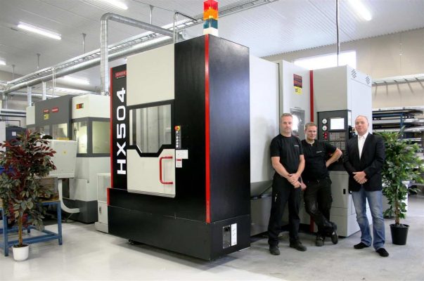 Tre män står bredvid en stor industrimaskin märkt "HX504" i fabriksmiljö med växter och rör i bakgrunden.