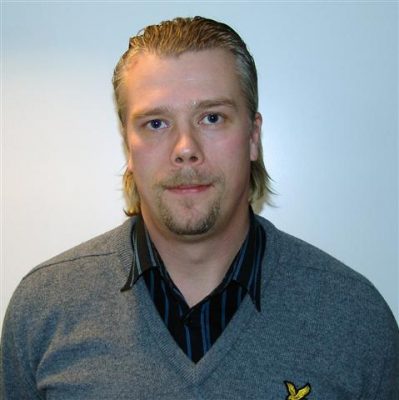En man med blont hår, kort bockskägg och mustasch, klädd i en grå tröja över en svart och blårandig skjorta, står vänd mot kameran mot en vanlig vit bakgrund.