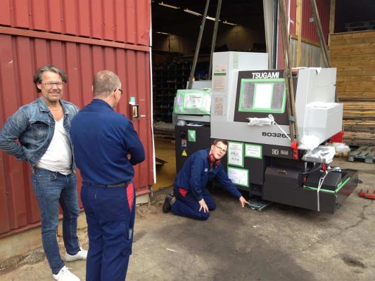 Tre män på en verkstad som inspekterar en ny tsugami b0326-maskin; en hukar vid basen, en annan diskuterar med en tekniker som står bredvid.