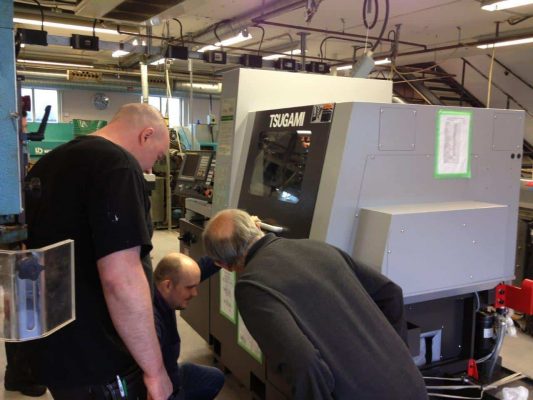 Tre män i en verkstad undersöker en tsugami cnc-maskin, till synes felsöker eller lär sig om dess funktion.