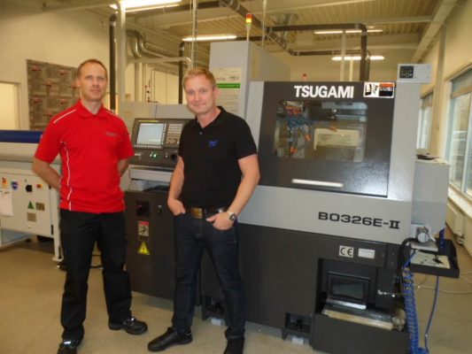 Två män står bredvid en tsugami b0326e-ii precisionsmaskin i en industriell miljö. en man har en röd skjorta, den andra i svart.