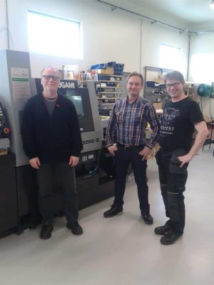Tre män står i en industriverkstad bredvid en stor maskin och ler mot kameran.