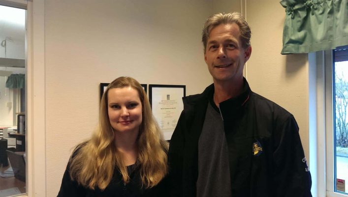 En man och en kvinna står på ett kontor och ler mot kameran med ett fönster och certifikat i bakgrunden.