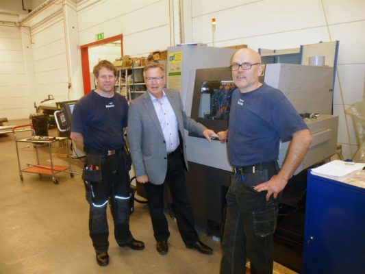 Tre män står i en verkstad, två iklädda arbetsoveraller och en i kostym, bredvid en stor industrimaskin.