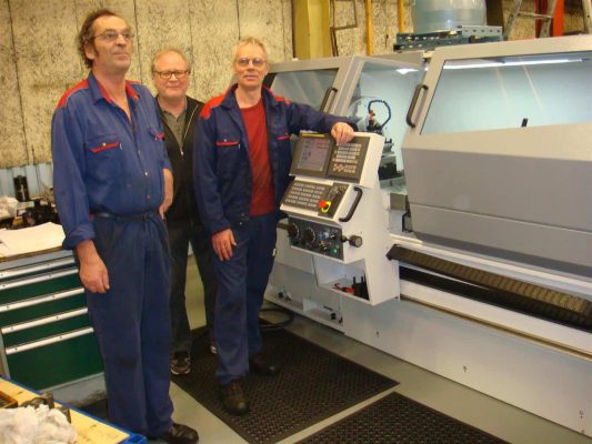 Tre arbetare i blå overaller som står bredvid en stor industrimaskin i en fabrik.