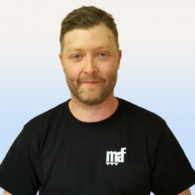 En man med ljusa skäggstubbar som ler mot kameran, klädd i en svart t-shirt med en "mf"-logotyp, mot en blå och vit bakgrund.