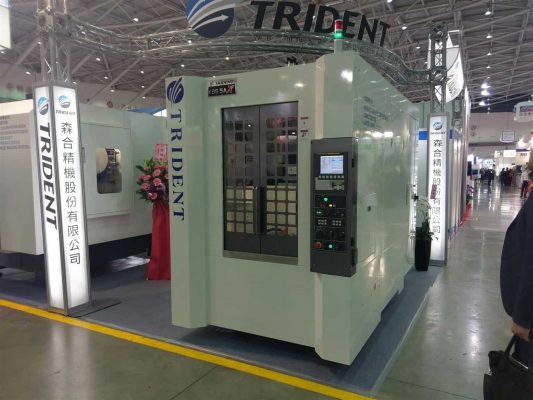 En stor industriell varuautomat märkt "trident" som visas på en mässa, med olika produkter synliga bakom en glaspanel.