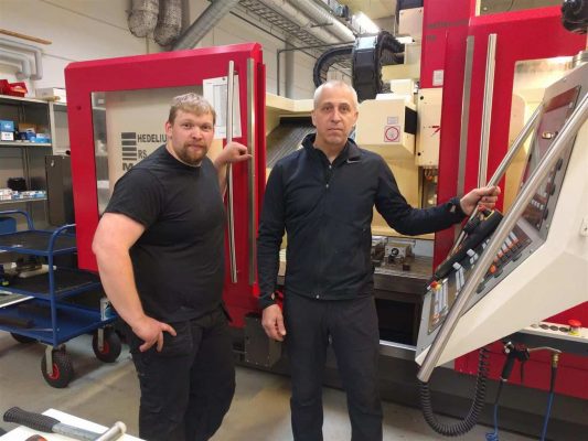 Två män som står bredvid en stor röd industrimaskin i en verkstadsmiljö.