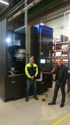 Två män som står bredvid en stor industriell cnc-maskin i en fabriksmiljö, en i en gul synlig jacka och den andra i en mörk kavaj.