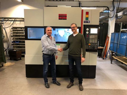Två män skakar hand framför en industrimaskin i en verkstadsmiljö.