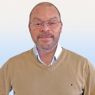 En medelålders man med glasögon och skägg ler mot kameran, klädd i en brun tröja över en vit skjorta mot en ljusblå bakgrund.