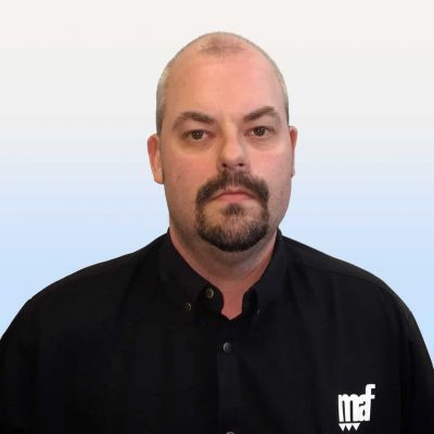 En medelålders kaukasisk man med kort skägg och mustasch, klädd i en svart skjorta med "mf"-logotyp, mot en lutning blå och vit bakgrund.