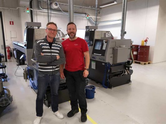 Två män som står i en verkstad med industrimaskineri i bakgrunden; den ena bär en randmönstrad jacka och jeans, och den andra bär en röd skjorta och svarta byxor.