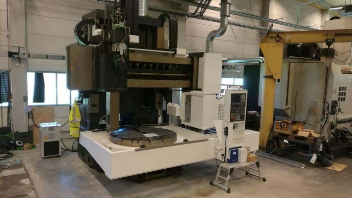 En stor industriell CNC-maskin är stationerad i en verkstad, omgiven av olika verktyg och utrustning. Maskinen har en kontrollpanel och en roterande plattform.