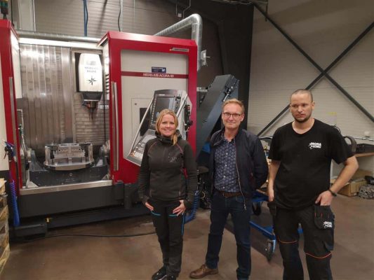 Tre personer som står framför en stor röd och silver industrimaskin i en fabriksmiljö.