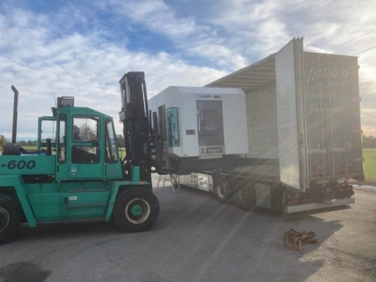 En stor grön gaffeltruck lastar av en Tornos-maskin från en semi-truck trailer under en delvis molnig himmel.