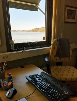 Ett hemmakontor med skrivbord, kringutrustning och en stol som vetter mot ett fönster med utsikt över vattnet. Vi söker vår nya ekonomiansvarige.
