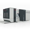 En stor, industriell CNC-maskin står på en plattform med kontrollpanel och trappsteg. Två förenklade humanoida figurer illustreras i närheten för skalajämförelse.