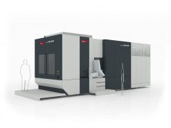 En stor, industriell CNC-maskin står på en plattform med kontrollpanel och trappsteg. Två förenklade humanoida figurer illustreras i närheten för skalajämförelse.