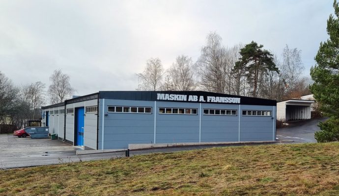En industribyggnad med skylten "maskin ab a. fransson" annonserar "Öppet Hus 25 maj - invigning av tillbyggnad" under mulen himmel.