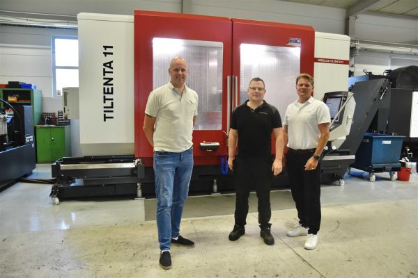 Tre män står i en industrianläggning med en maskin märkt "Hedelius Tiltenta 11" i bakgrunden.