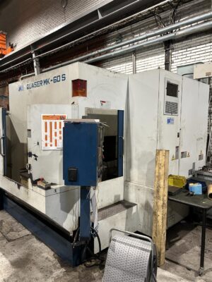 En stor Quaser MK-60S CNC-bearbetningscentral står i en industriell verkstad. Papper är tejpade på maskinen och verktyg och material ligger utspridda på ett bord i närheten. Området har synliga tegelväggar och rörledningar.