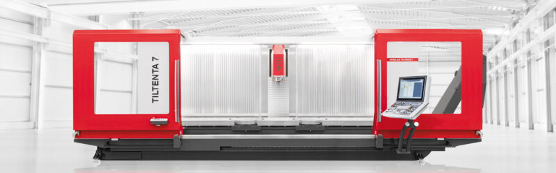 En stor rödvit industriell CNC-maskin märkt "TILTENTA 7" står i en ljus, rymlig och modern fabrik med vita väggar och takbjälkar. Maskinen har en sluten arbetsyta och en kontrollpanel med bildskärm.